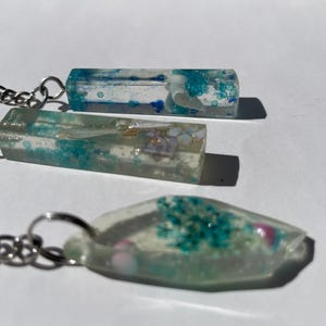 May include: Three clear resin keychains with silver-colored chains. The keychains contain dried flowers and blue and white accents. The top two keychains are rectangular, and the bottom one is oval-shaped.