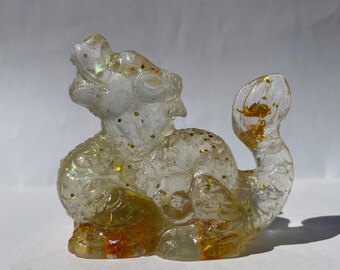 May include: A clear resin figurine of a mythical creature with gold glitter accents. The figurine has a detailed design, with a dragon-like head and a curled tail. The base has a golden-brown hue.
