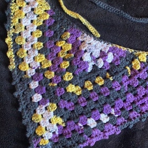 May include: A crocheted triangle scarf in black, purple, yellow, and white. The scarf is made with a granny square stitch.