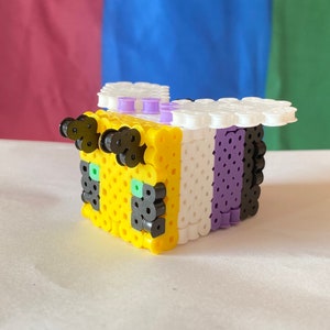 May include: A yellow, white, and purple beaded bee with black eyes and green accents. The bee is made of small plastic beads and is a popular craft project.