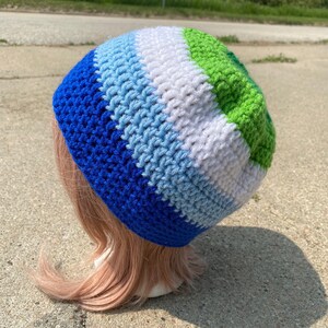 May include: A crocheted beanie hat with horizontal stripes in shades of blue, light blue, white, and green. The hat is handmade and appears to be soft and comfortable. The hat is shown on a mannequin head.
