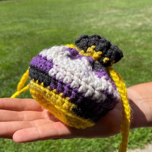May include: A crocheted pouch with a yellow, black, white, and purple color scheme. The pouch has a drawstring closure and a long yellow cord.