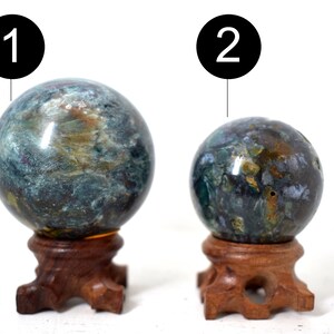 Puede incluir: Dos esferas de ágata verde, marrón y dorada sobre soportes de madera. Las esferas están pulidas y tienen un patrón de remolino. La esfera más grande está etiquetada con "1" y la esfera más pequeña está etiquetada con "2".