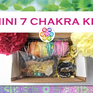May include: A small cardboard box filled with items for a chakra kit. The box contains a white sage smudge stick, a beaded bracelet, a clear quartz crystal, a black stone with the word "Happiness" on it, and a small bag of colourful stones. The box is decorated with yellow shredded paper and two carnations, one pink and one yellow.
