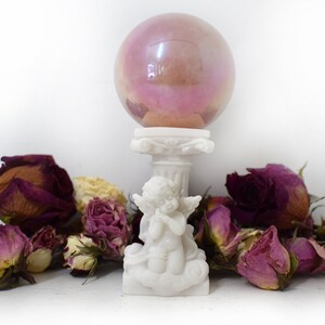 K&ouml;nnte beinhalten: Eine wei&szlig;e Cherubstatue, die eine rosafarbene, schillernde Glaskugel auf einem Sockel h&auml;lt, umgeben von getrockneten Rosen.