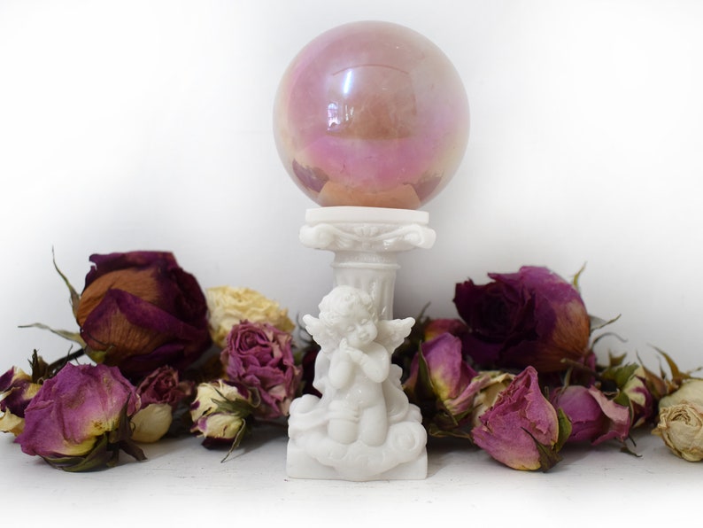 K&ouml;nnte beinhalten: Eine wei&szlig;e Cherubstatue, die eine rosafarbene, schillernde Glaskugel auf einem Sockel h&auml;lt, umgeben von getrockneten Rosen.