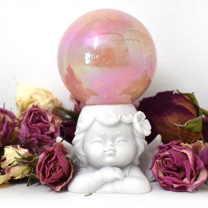 K&ouml;nnte beinhalten: Eine wei&szlig;e Porzellan-Cherubfigur mit Fl&uuml;geln, die eine rosafarbene, irisierende Glaskugel auf dem Kopf h&auml;lt. Der Cherub ist von getrockneten rosa Rosen umgeben.