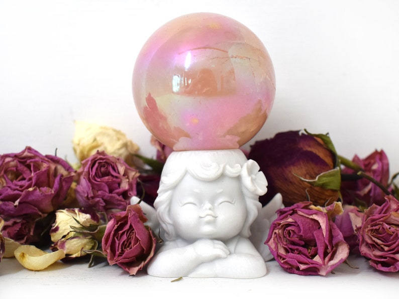 K&ouml;nnte beinhalten: Eine wei&szlig;e Porzellan-Cherubfigur mit Fl&uuml;geln, die eine rosafarbene, irisierende Glaskugel auf dem Kopf h&auml;lt. Der Cherub ist von getrockneten rosa Rosen umgeben.