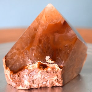 Puede incluir: Un gran cristal de ágata naranja claro con una base natural rugosa. El cristal tiene forma de pirámide y tiene una apariencia translúcida y vidriosa.