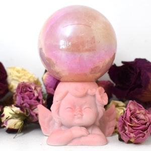 K&ouml;nnte beinhalten: Eine rosafarbene Cherubfigur, die eine rosafarbene, irisierende Glaskugel h&auml;lt. Die Figur ist von getrockneten Rosen umgeben.