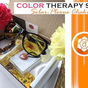 Puede incluir: Un juego de caja de regalo para terapia de color, con un tema de chakra del plexo solar. El juego incluye un par de gafas de sol teñidas de ámbar, un brazalete de ojo de tigre, un colgante de corazón de cornalina y una pequeña tarjeta con la palabra "Citrino" en ella. La caja está decorada con papel triturado amarillo y la tapa tiene las palabras "COLOR THERAPY SET Solar-Plexus Chakra" impresas en ella.