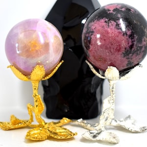 May include: Two crystal spheres on decorative stands. The sphere on the left is a light pink and purple colour. The sphere on the right is a dark pink and black colour. Both stands are gold and silver.