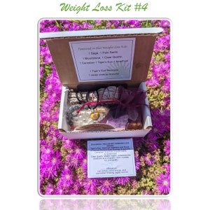 May include: A white box with a green and white label that reads "Weight Loss Kit #4" and lists the contents: 1 Sage, 1 Palo Santo, 1 Bloodstone, 1 Clear Quartz, 1 Carnelian, 1 Tiger's Eye, 1 Amethyst, 1 Tiger's Eye Necklace, 1 seven chakras bracelet. The box is open and contains various crystals, a sage bundle, and a red string bracelet with a tiger's eye pendant. The box is sitting on a bed of pink flowers.