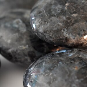 Puede incluir: Primer plano de un grupo de piedras pulidas lisas, grises y negras. Las piedras tienen un brillo sutil y una calidad ligeramente iridiscente.