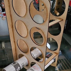 Puede incluir: Un estante de almacenamiento de madera con cortes ovalados, diseñado para guardar materiales enrollados. Se ven rollos de vinilo blanco y negro dentro del estante. El estante es de madera marrón claro.