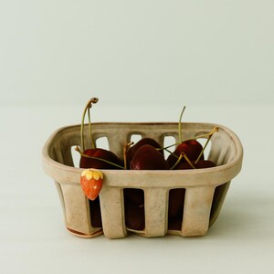 May include: A small, beige ceramic basket filled with ripe, dark red cherries. The basket has a rectangular shape with vertical slots and a decorative strawberry charm. The cherries have green stems.