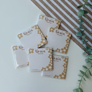 Op de afbeelding: Zes witte plakbriefjes met een bruine bloemenrand en de tekst "Brain Dump" in bruine letters. De briefjes liggen op een wit oppervlak met een bruin-wit gestreepte stof op de achtergrond.