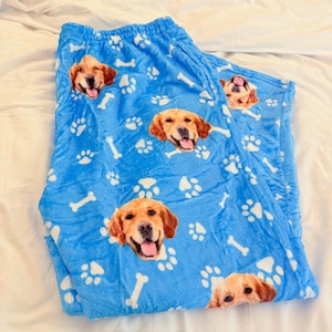 May include: Blue pajama pants with a repeating pattern of golden retriever faces, white paw prints, and bone shapes. The pants are made of a soft, plush material.