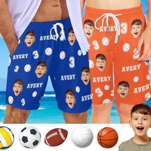 May include: Two pairs of blue and orange swim trunks with a baseball theme. The trunks feature a repeated pattern of baseballs, the number "3", and a person's face. The text "Avery" is repeated on the trunks.