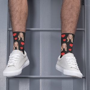 May include: A pair of black socks with a red heart pattern and a woman's face printed on them. The socks are being worn by a person with white sneakers.