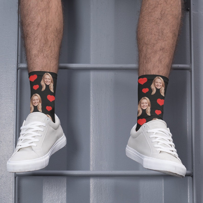 May include: A pair of black socks with a red heart pattern and a woman's face printed on them. The socks are being worn by a person with white sneakers.