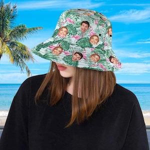 May include: A bucket hat featuring a repeating pattern of green tropical palm leaves, pink flamingos, and multiple small portraits of a person's face. The hat has a wide, downward-sloping brim and a rounded crown, constructed from fabric.