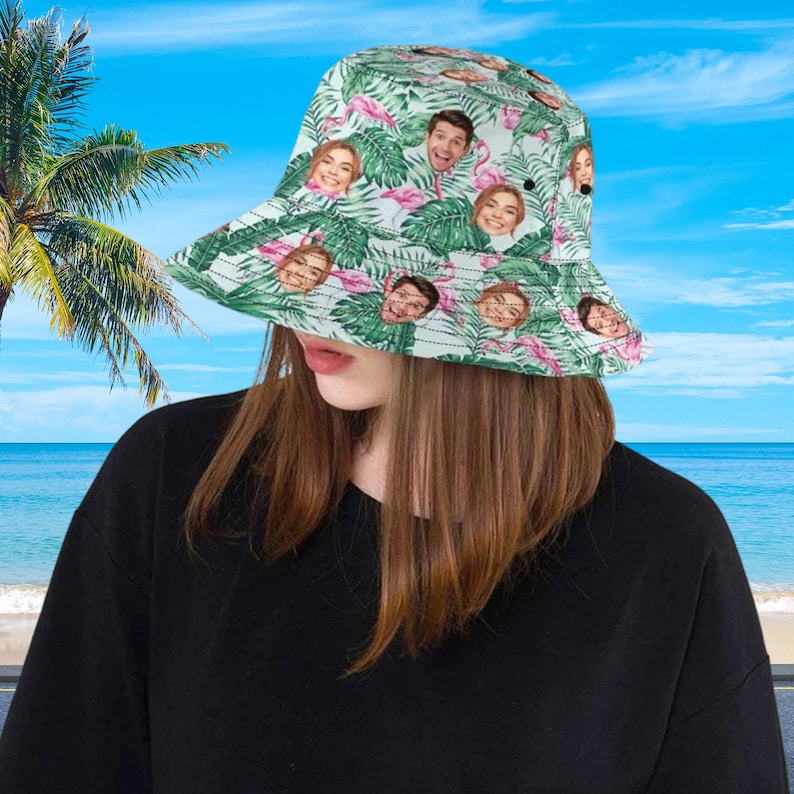 May include: A bucket hat featuring a repeating pattern of green tropical palm leaves, pink flamingos, and multiple small portraits of a person's face. The hat has a wide, downward-sloping brim and a rounded crown, constructed from fabric.
