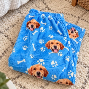 May include: Bright blue fleece pajama pants featuring a repeating pattern of golden retriever dog faces, white paw prints, and white bones. The soft fabric has an elastic waistband and a relaxed fit, showcasing a vibrant canine-themed design.