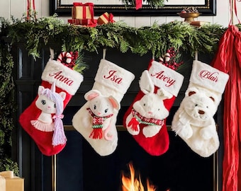 Op de afbeelding: Vier kerstsokken hangen aan een schoorsteenmantel. De sokken zijn voorzien van pluche dierenfiguren: een eenhoorn, een muis, een konijn en een beer. Elke witte boord is geborduurd met de namen Anna, Isa, Avery en Blair in rode letters.