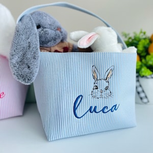 May include: A blue and white striped fabric tote bag with a bunny rabbit face and the name "Luca" embroidered on the front. The bag is filled with stuffed animals.