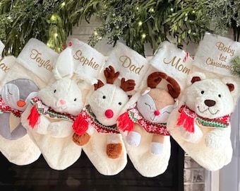 Op de afbeelding: Een rij van zes crèmekleurige pluche kerstsokken die op een rij hangen. Elke sok is voorzien van een ander knuffeldier met een rood, groen en wit patroon sjaaltje. De namen Max, Taylor, Sophie, Dad, Mom en Baby 1st Christmas zijn in gouddraad op de boorden geborduurd.