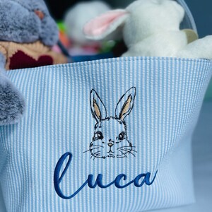 May include: Blue and white striped fabric storage basket with a bunny embroidered on the front. The basket is personalized with the name "Luca" in blue embroidery.