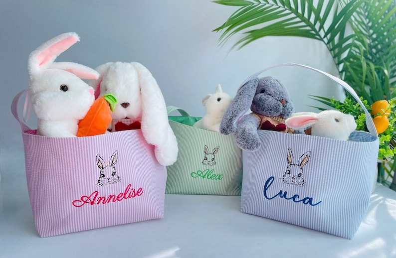 May include: Three striped fabric Easter baskets with bunny ears handles. Each basket is filled with stuffed animals and has a name embroidered on the front: Annelise, Alex, and Luca.