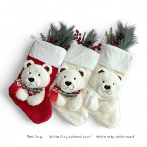 May include: Three Christmas stockings with polar bear faces. One stocking is red, and the other two are white. Each bear wears a scarf. The stockings are decorated with faux pine and berries.