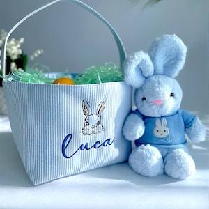 May include: A blue and white striped Easter basket with a bunny embroidered on the front. The basket is filled with green grass and has a blue handle. A blue plush bunny with a blue shirt is sitting next to the basket. The basket is personalized with the name "Luca".