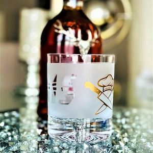 May include: A frosted glass with a gold design of a chef's hat and crossed knives. The glass is sitting on a table with a bottle of liquor in the background.
