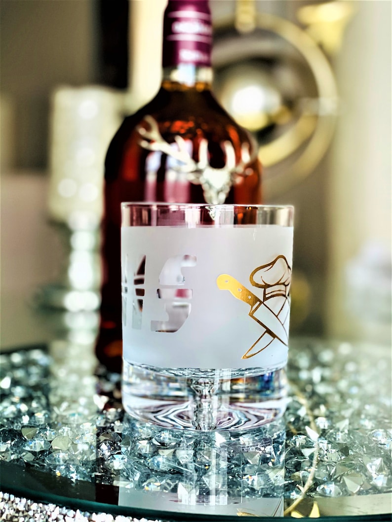 May include: A frosted glass with a gold design of a chef's hat and crossed knives. The glass is sitting on a table with a bottle of liquor in the background.