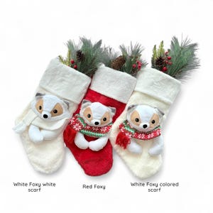 May include: Three Christmas stockings with fox ornaments. One stocking is red, and two are white. Each fox wears a scarf. The stockings are decorated with pine and berries.