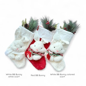 May include: Three Christmas stockings featuring bunny designs. Two are white with white scarves, and one is red with a patterned scarf. Each stocking has a plush bunny face and is decorated with greenery and berries.