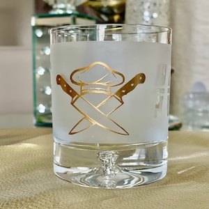 May include: A frosted glass with a gold chef hat and crossed knives design. The glass has a clear base.