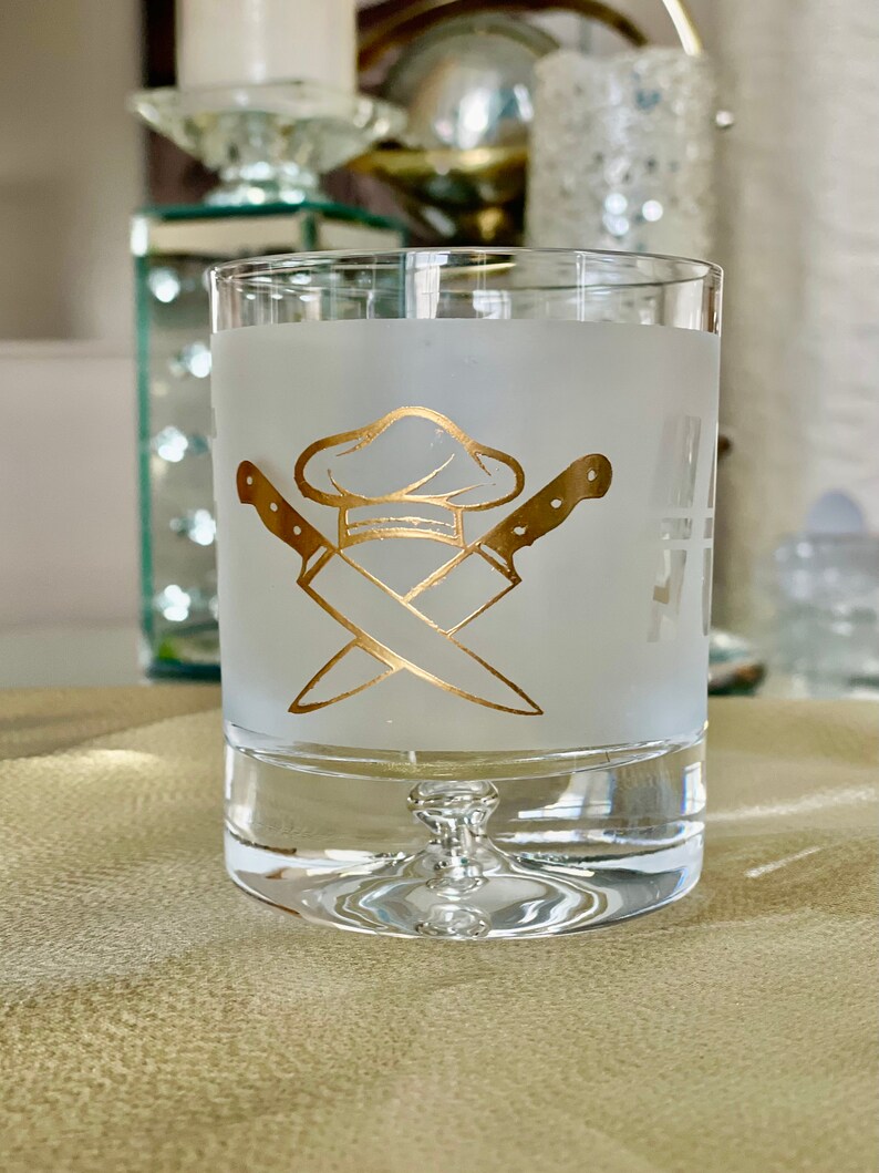 May include: A frosted glass with a gold chef hat and crossed knives design. The glass has a clear base.