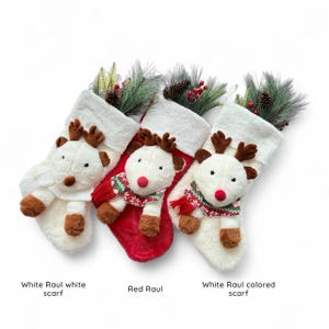 May include: Three Christmas stockings with reindeer faces and plush bodies. One stocking is red, and the other two are white. Each reindeer has brown antlers, a red nose, and a scarf. The stockings are decorated with pine needles, berries, and pine cones.