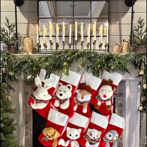 May include: A festive Christmas fireplace display. Red and white stockings with plush animal characters hang from the mantel. The mantel is decorated with greenery, candles, and a large mirror.