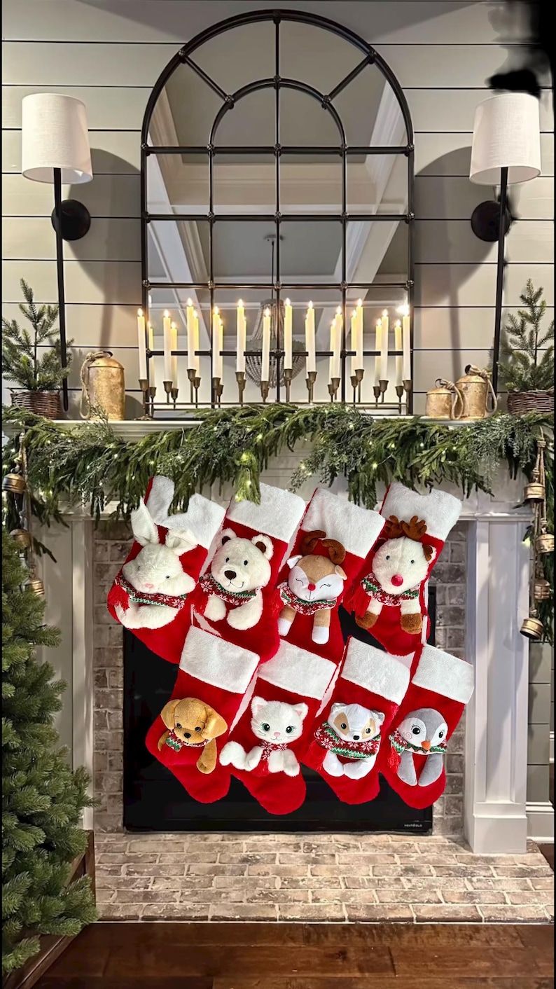 May include: A festive Christmas fireplace display. Red and white stockings with plush animal characters hang from the mantel. The mantel is decorated with greenery, candles, and a large mirror.