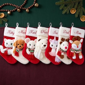 Könnte beinhalten: Sieben Weihnachtsstrümpfe mit weißen Kunstfellbündchen, bestickt mit den Namen Kitty, Max, Karl, Blair, Isa, Dad und Mom. Jeder Strumpf zeigt eine Plüschtierfigur, darunter eine Katze, ein Hund, eine Maus, ein Bär, ein Einhorn, ein Rentier und ein Reh.