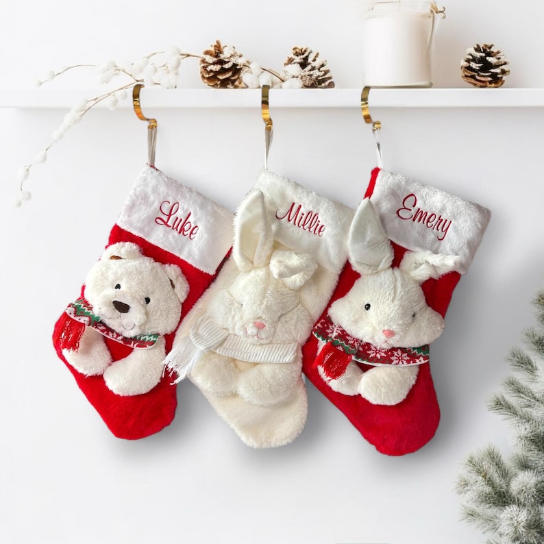 May include: Three Christmas stockings with names embroidered in red. One features a white teddy bear, and two feature white bunny rabbits. All are red with white faux fur trim and gold hanging hooks.