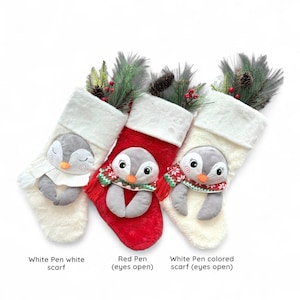 May include: Three Christmas stockings featuring penguin designs. One is red, and two are white. Each stocking has a plush penguin face and is decorated with greenery and pine cones. The text at the bottom reads "White Pen white scarf", "Red Pen (eyes open)", and "White Pen colored scarf (eyes open)".