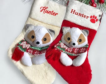 Op de afbeelding: Twee kerstsokken, één crème en één rood, elk met een pluchen hondenspeeltje met een feestelijke sjaal. De crèmesok heeft "Hunter" in het rood geborduurd. De rode sok heeft "Hunter" en een pootafdruk.