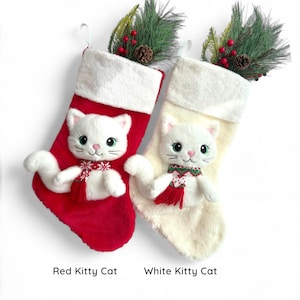 May include: Two festive Christmas stockings, one red and one cream, each featuring a white cat with a scarf. The stockings have faux fur trim and are decorated with greenery. Text at the bottom reads "Red Kitty Cat" and "White Kitty Cat."