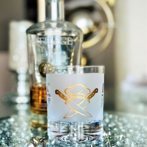 May include: A frosted glass with a gold chef hat and crossed knives design. The glass is sitting on a table with a bottle of liquor in the background.